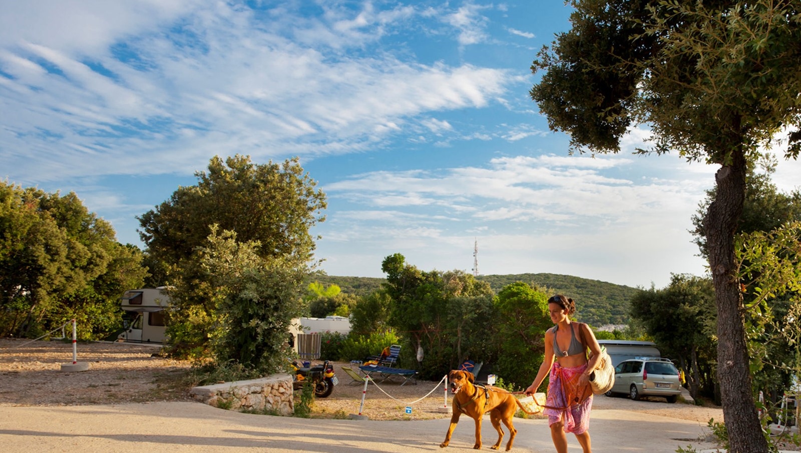 Eine Person geht mit einem Hund an einem sonnigen Tag in der Nähe eines Campingplatzes auf Cres spazieren